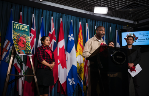 A man in brown clothes stands at a podium speaking with two women flanked at his sides and the flags of Canadian provinces behind him.