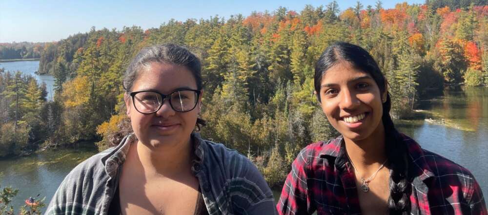 Julie-Ann Arbour (left) and Gurleen Chana take a selfie in front of lakes and a forest in the fall.