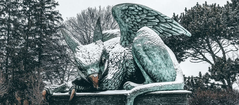 A copper statue of a gryphon covered in snow in front of trees.