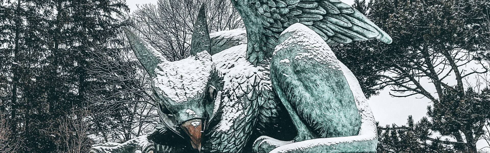 A copper statue of a gryphon covered in snow in front of trees.