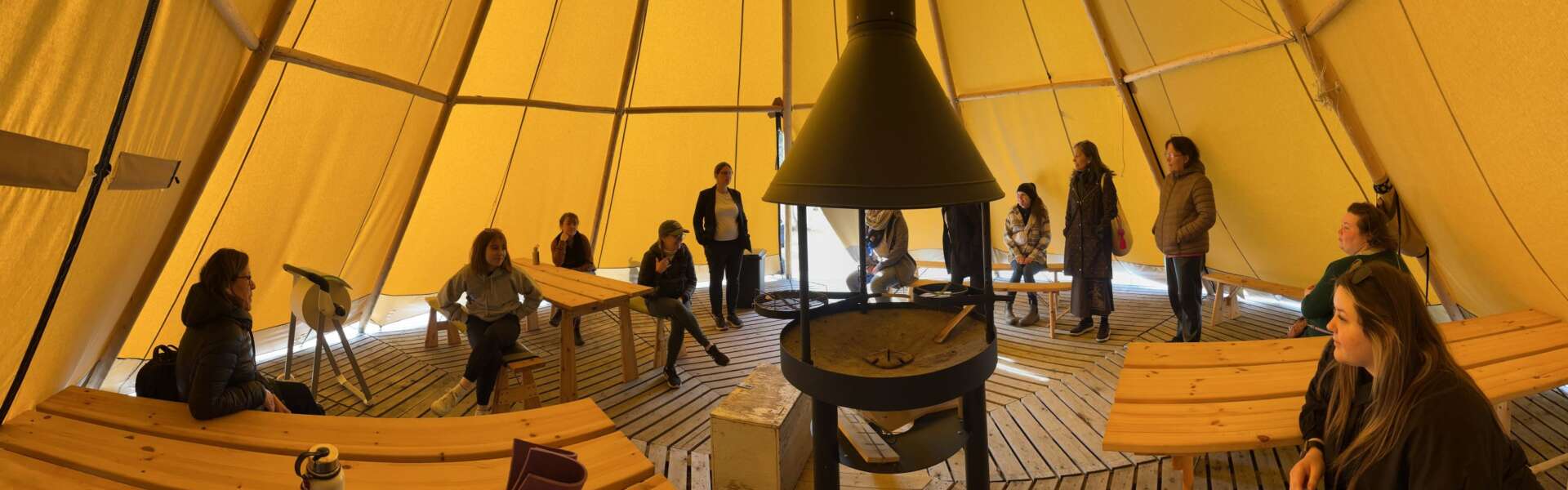People sit on wooden benches inside a circular tent with a woodstove in the middle.