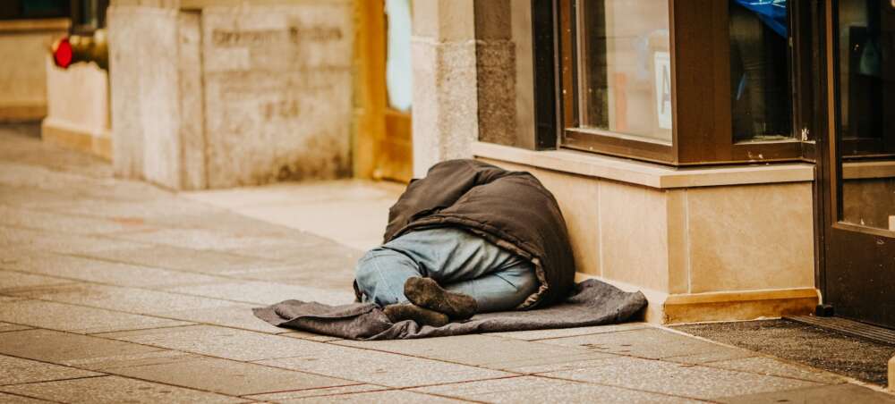 A person in a coat, jeans and black socks lies on a sidewalk in front of a store.