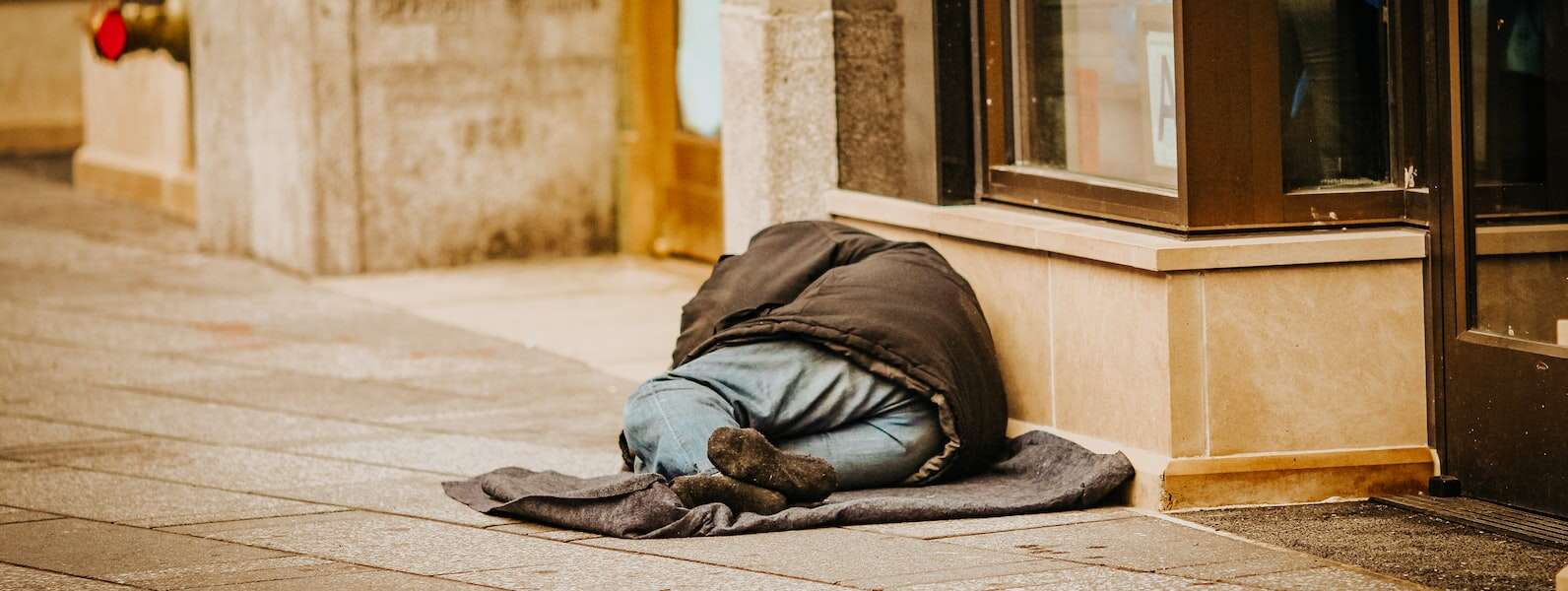 A person in a coat, jeans and black socks lies on a sidewalk in front of a store.