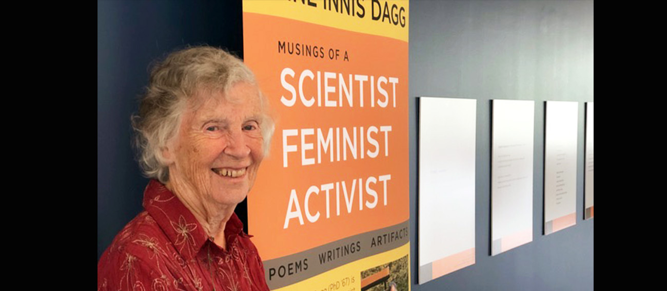 Anne Innis Dagg stands beside a wall displaying some exhibit items