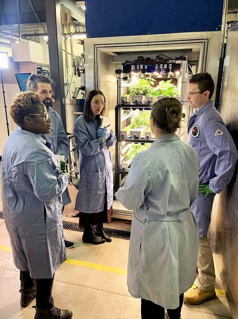Four people in lab coats as a fifth person speaks in front of a large refrigerator-sized chamber filled with trays of growing vegetables
