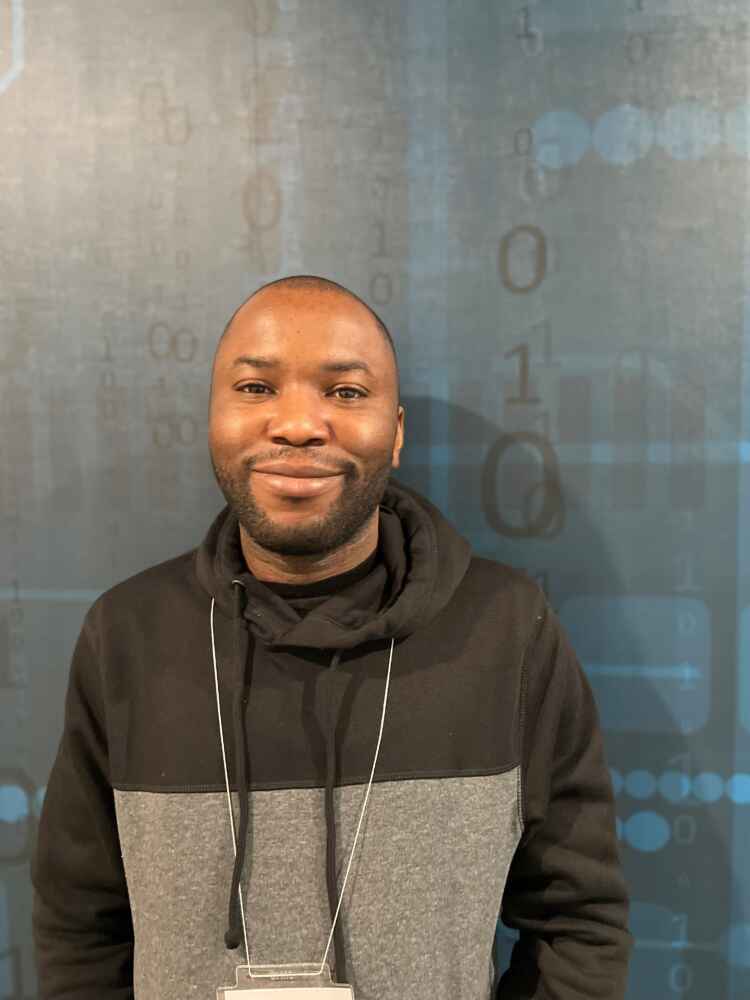 Charles Obi, current MCTI international graduate student, is smiling for the camera, standing in front of a wall displaying binary numbers