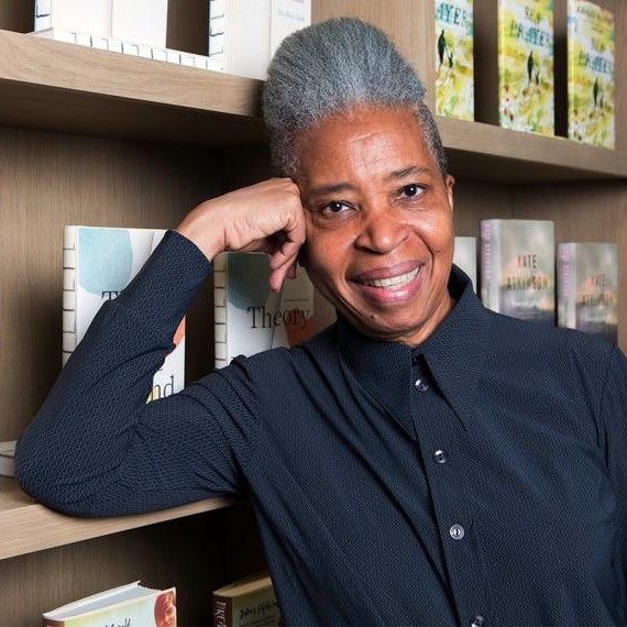 Dionne Brand smiles while leaning against a bookshelf