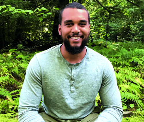 Kai Thomas in grey long-sleeve shirt is seated in front of greenery, smiling and leaning slightly forward with his arms resting on his knees.