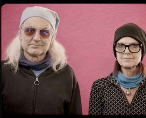 A person with long gray hair, head scarf and sunglasses stands in front of pink background beside a person with black toque, glasses, scarf and buttoned shirt. 