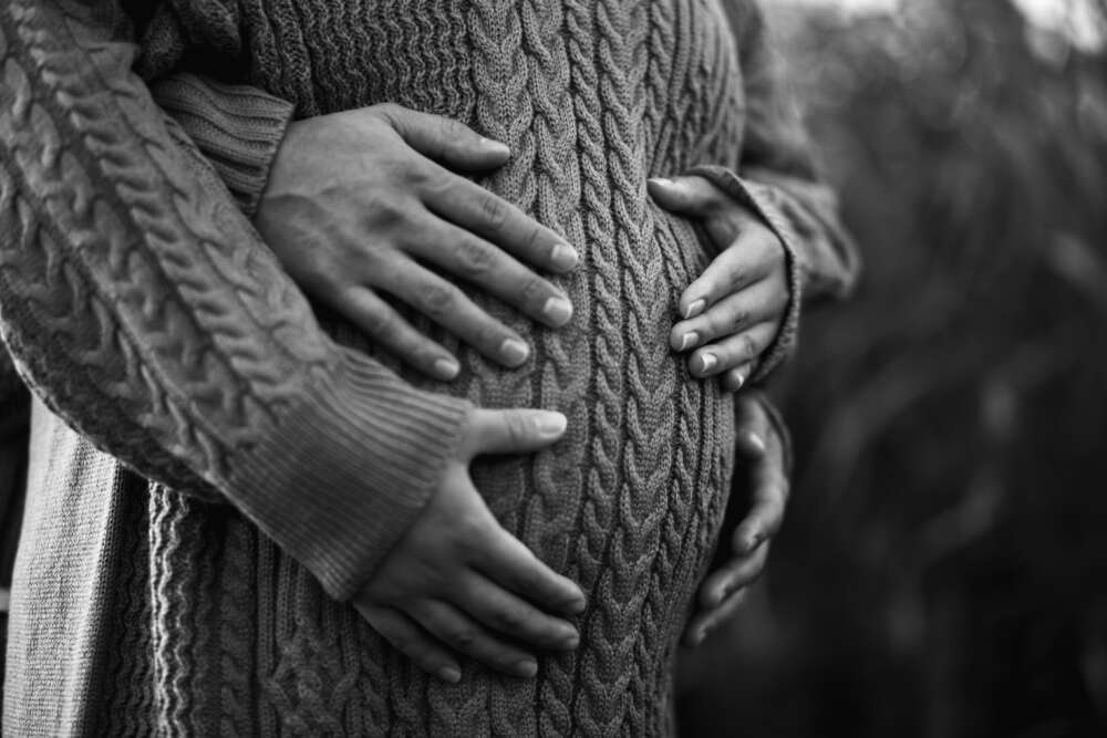 A black and white photo of a person's pregnant belly is cupped by their and their partners hands. The pregnant person is wearing a cable knit dress.
