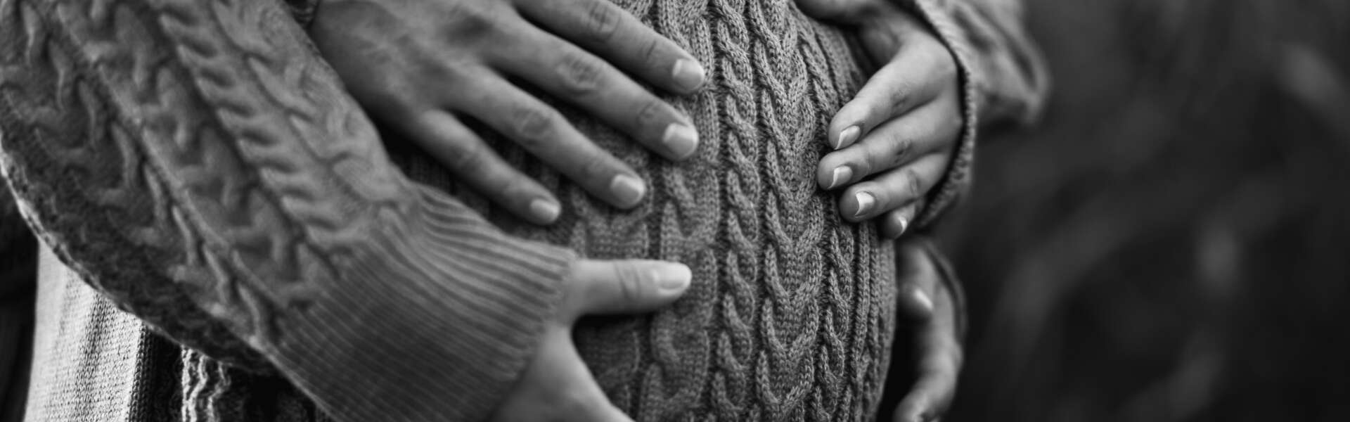 A black and white photo of a person's pregnant belly is cupped by their and their partners hands. The pregnant person is wearing a cable knit dress.