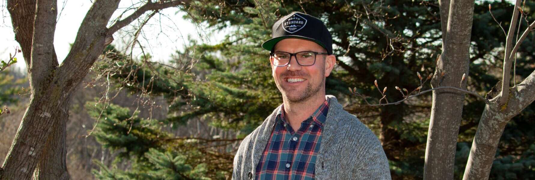 A person stands in front of evergreen trees, facing the camera, smiling, wearing a black baseball hat, black-framed glasses, a plaid shirt and a grey sweater.