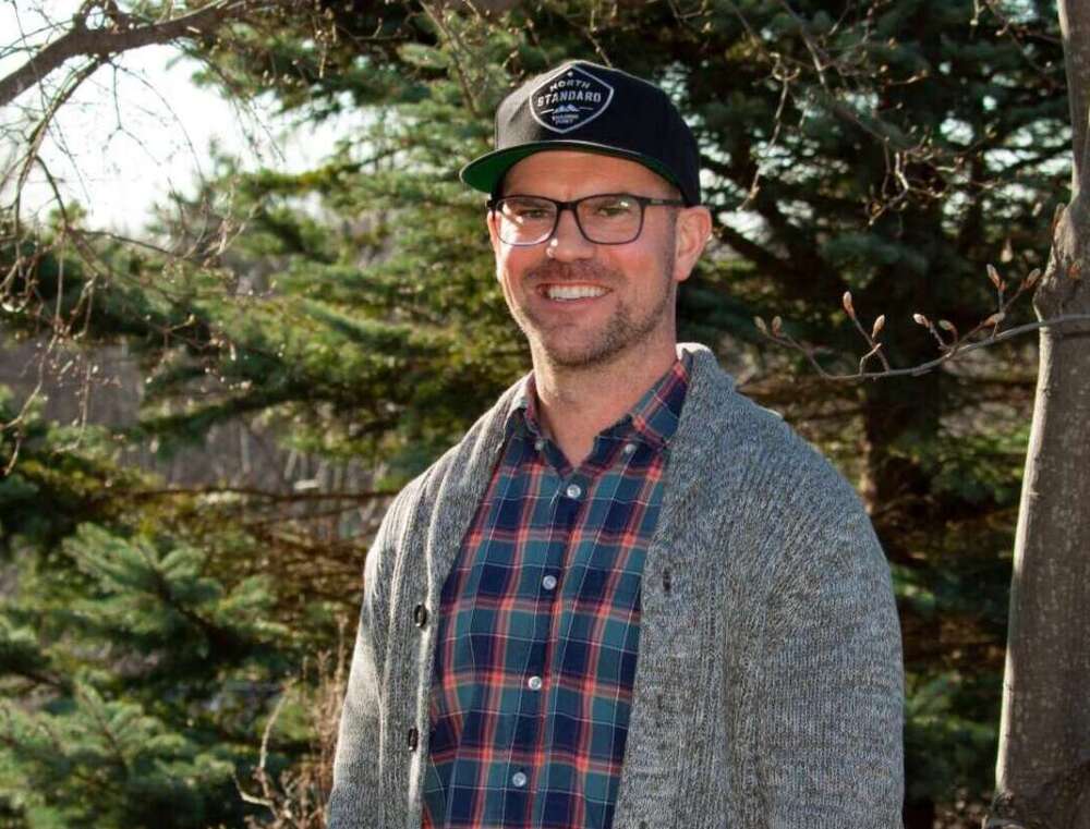 A person stands in front of evergreen trees, facing the camera, smiling, wearing a black baseball hat, black-framed glasses, a plaid shirt and a grey sweater.