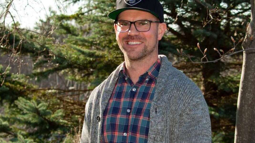 A person stands in front of evergreen trees, facing the camera, smiling, wearing a black baseball hat, black-framed glasses, a plaid shirt and a grey sweater.