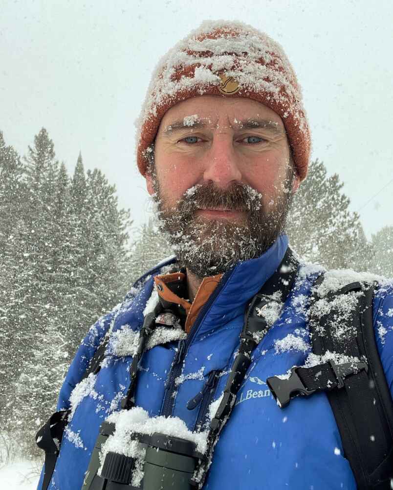 A man wears binoculars around his neck while standing in a snowy forest