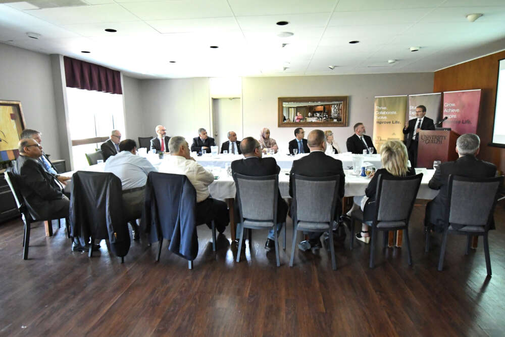 Several people sit around a U-shaped boardroom table while a person speaks at a podium