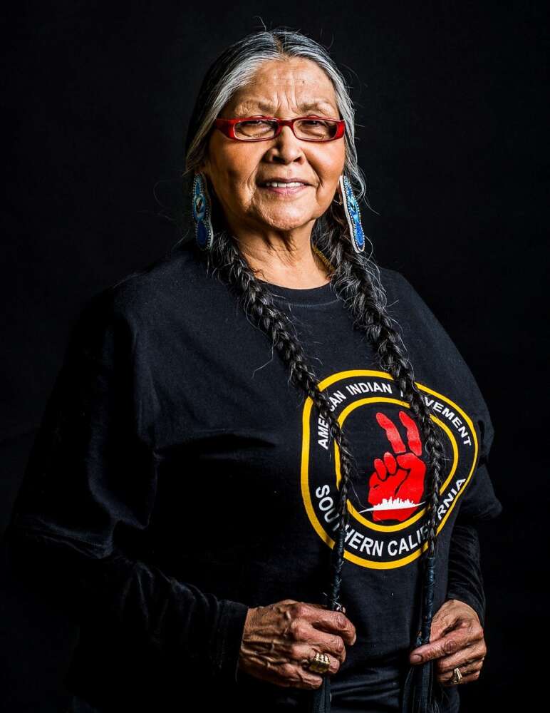 Mona Stonefish, Bear Clan, Ogichidaakwe of the Anishinaabek Three Fires Confederacy, wears a black t-shirt that depicts a red hand giving the peace sign and reads "American Indian Movement Southern California." She is smiling and her hands are holding her two long braids as she stands