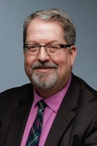 A man with brown hair, glasses and a beard smiles wearing a pink shirt, brown suit jacket and multi-coloured tie.