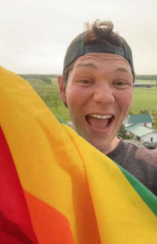 A man with a big smile wearing a green backwards ball cap stands with a red, orange, yellow and green Pride flag in his hand.