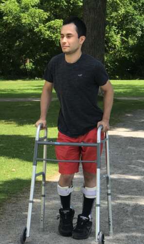 A brown-haired man in a dark gray t-shirt and red shorts smiles as he walks with the aid of a walker that his hands are holding onto.