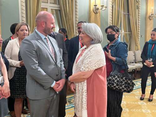 A man in with a bald head and beard in a gray suit stands with his hands clasped in front of him talking to a gray-haired woman in a pink dress and white shawl. 