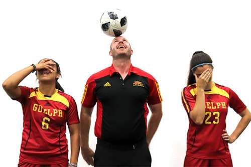 Two athletes in red and gold Gryphon soccer uniforms stand on either side of a person in a black Gryphon coaches shirt balancing a soccer ball on his head.