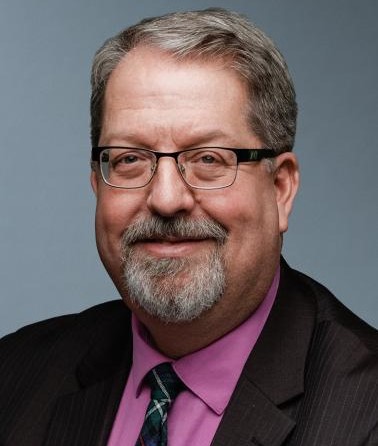 A man with brown hair, glasses and a beard smiles wearing a suit tie