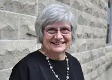 Dr. Paula Brauer stands against a grey brick wall, smiling, wearing glasses with a necklace over a black top.