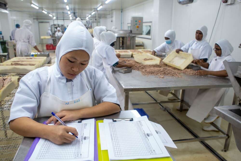 a person in a white protective gown and hood fills out food fastey paperwork in a warehouse with several food workers in teh background