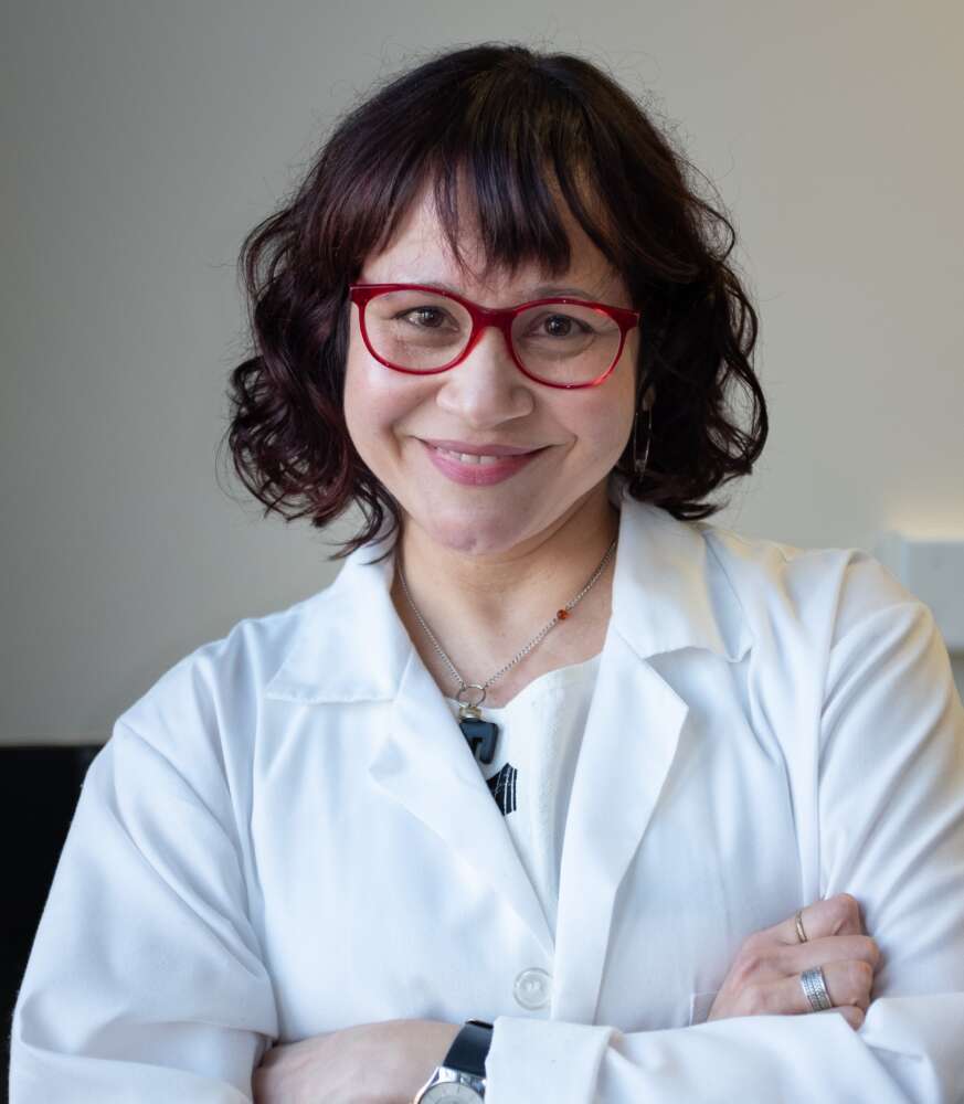 Dr. Alicia Viloria-Petit wears a lab coat and crosses their arms for a portrait