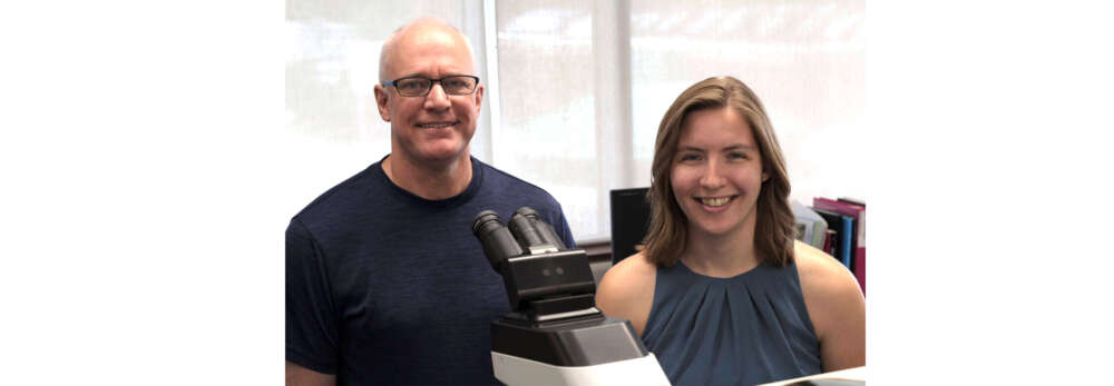 Two people sit near a microscope in a lab, smiling for the camera