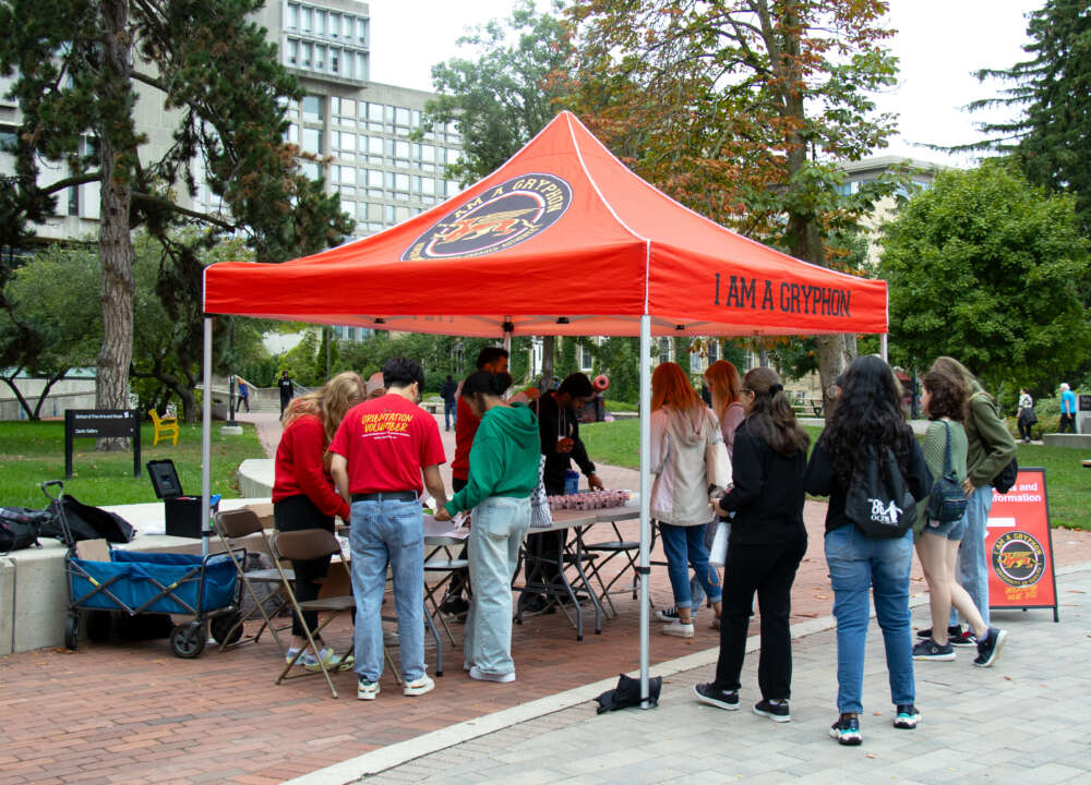 Students stop to speak to Orientation Volunteers under a large red tent with the words 'I am a Gryphon'