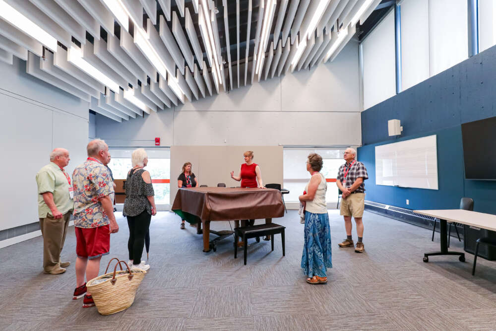 Several people stand around a grant piano in abright light filled rehearsal space