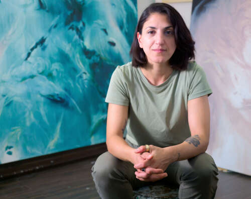 A person with shoulder-length dark brown hair sits with their elbows resting on their bent knees wearing a light green top and dark green pants in front of their blue and green painting.