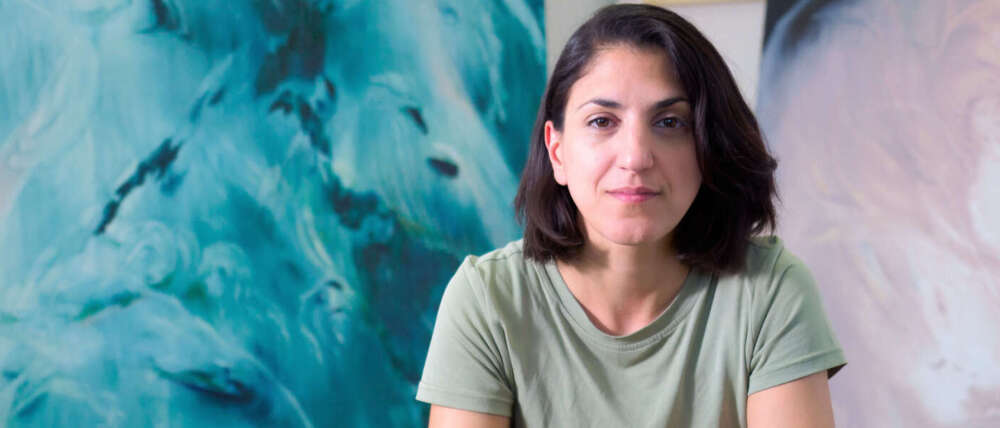 A person with shoulder-length dark brown hair sits with their elbows resting on their bent knees wearing a light green top and dark green pants in front of their blue and green painting.