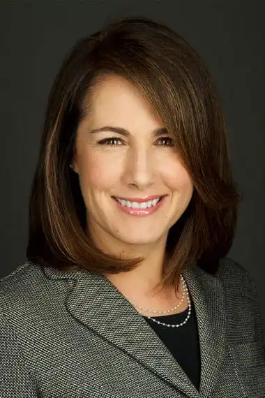 A person with shoulder-length brown hair smiles into the camera wearing a black top, gray blazer and two-strand necklace.