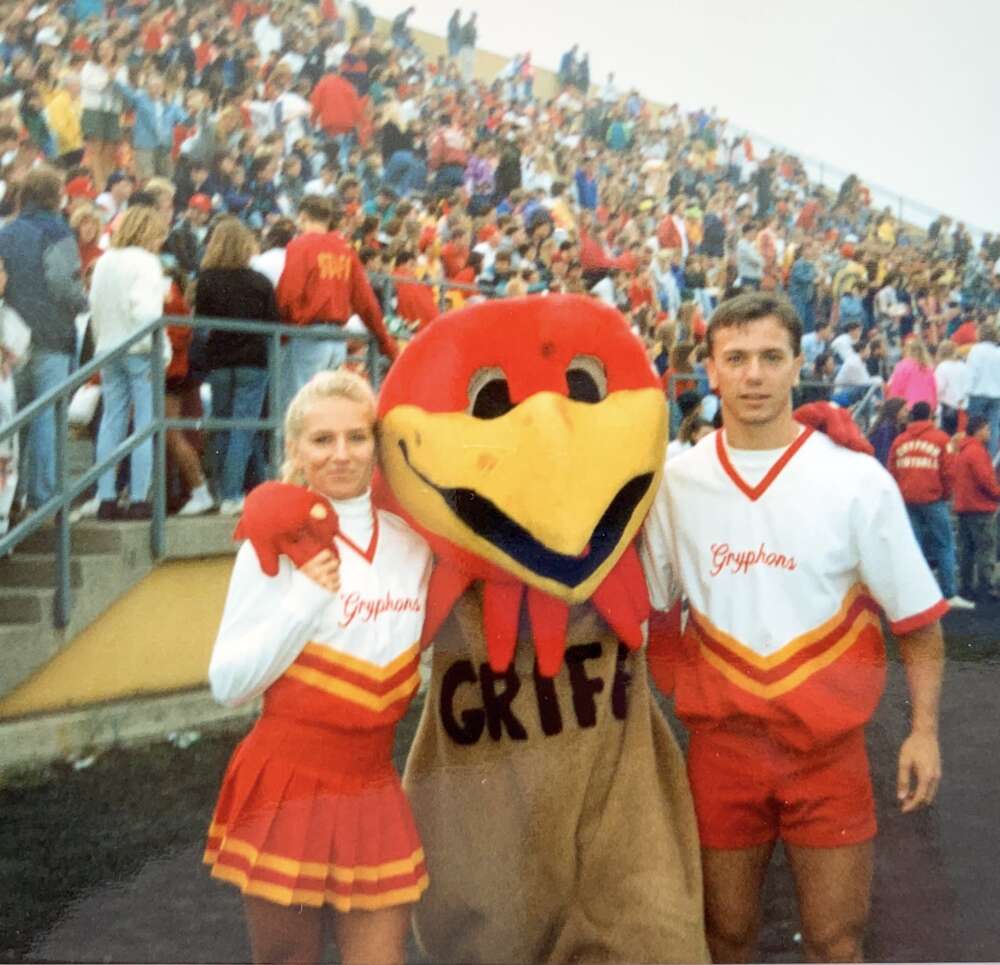 Two people in cheerleading costumes put thir arms around a large bird mascot before a crowd of people on bleachers
