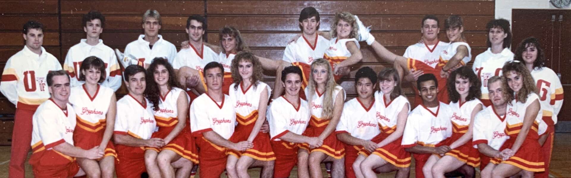 A group of 25 students in cheerleader uniforms pose for a portrait in a university gym
