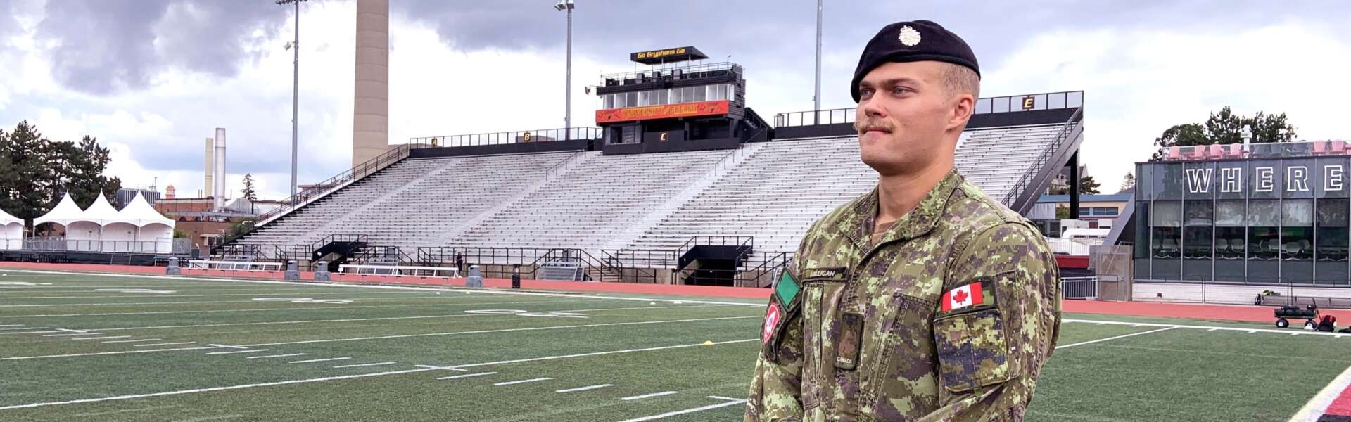 a reservist soldier wears an operational uniform while standing on a football field with bleachers behind