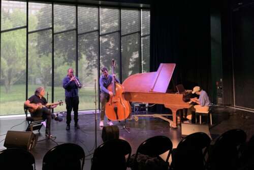 Four musicians including one on a grand piano play in a dark theatre with large windows looking out onto green space
