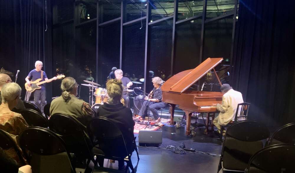 Four musicians including one on a grand piano play in a dark theatre with large windows behind