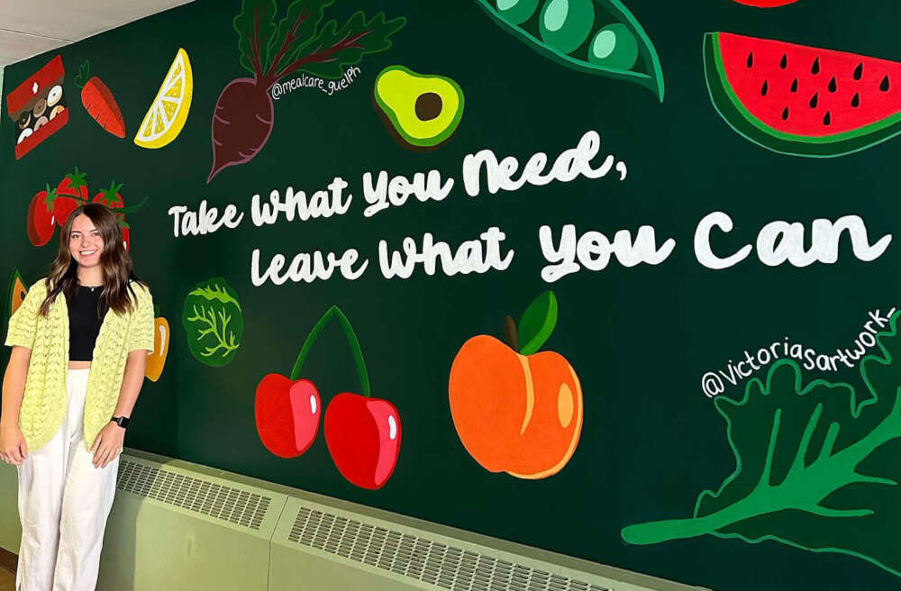 A person with long, wavy brown hair stands in a black top, yellow sweater, khaki pants and white shoes in front of a mural of red, orange, green and yellow fruits and vegetables with white text reading, take what you need, leave what you can.