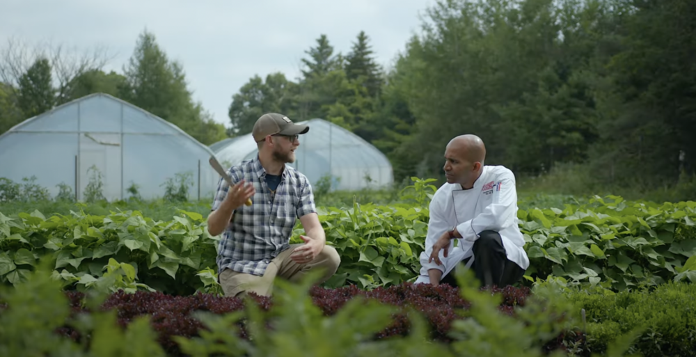 A man in a plaid shirt, a beard, glasses and a baseball cap holds a small shovel while crouched in a garden beside a second man dressed in chef whites.