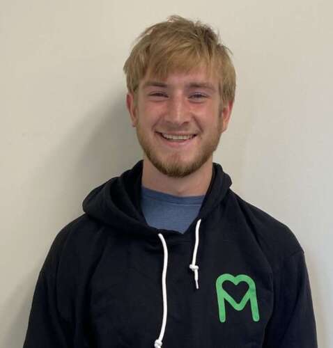 A person with short blond hair and a beard wearing a blue t-shirt under a black hoodie with a green MealCare logo stands smiling into the camera.