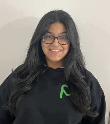 A person with long, brown hair and glasses stands in a black sweatshirt with a green MealCare logo smiling.