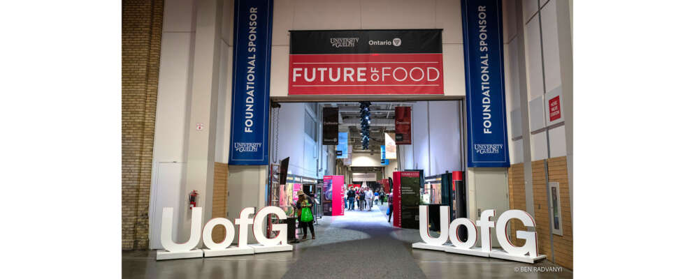 Blue and red signs with text that say University of Guelph are hung above a hall where people walk among exhibits showcasing research at the Royal Agricultural Winter Fair