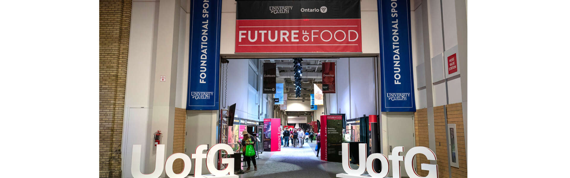 Blue and red signs with text that say University of Guelph are hung above a hall where people walk among exhibits showcasing research at the Royal Agricultural Winter Fair