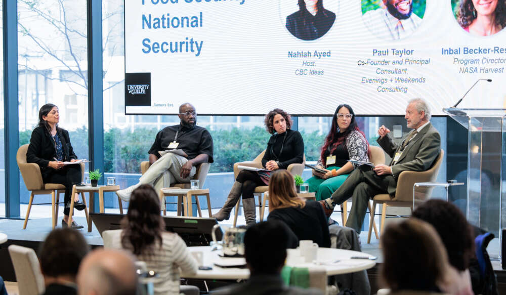 Five people set in chairs in front of a large screen while speaking to a room of participants