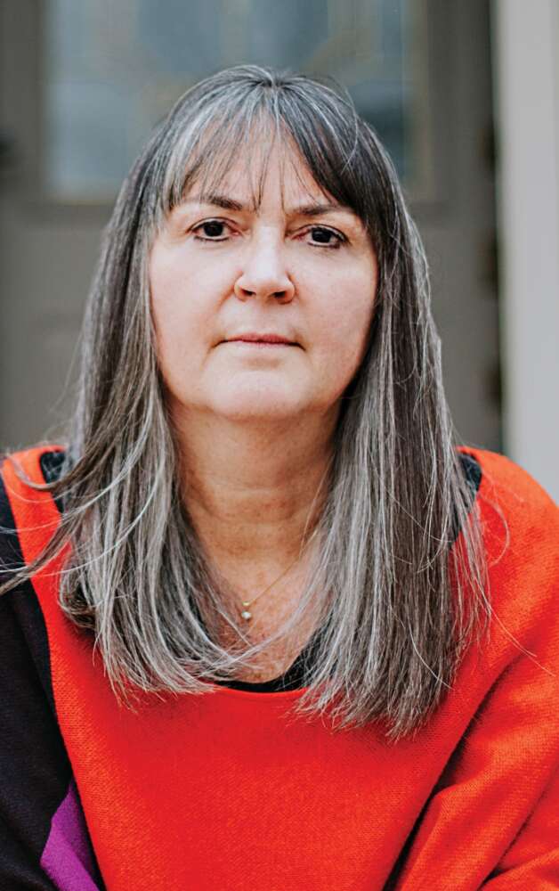A person with long, gray and brown hair with bangs wears a red sweater and looks into the camera in front of a gray stone building.