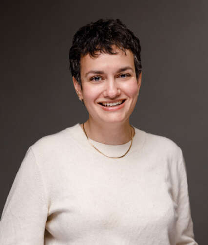 A person in a creme-coloured sweater with short brown hair stands against a charcoal background smiling into the camera